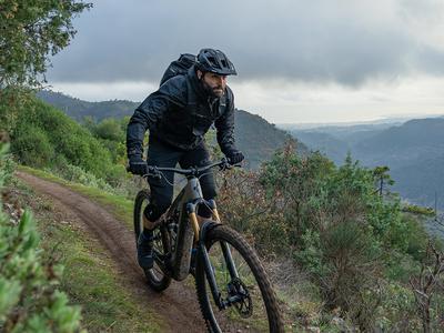 Mann fährt mit Cube E-Gravel-Bike auf einem Bergpfad, umgeben von Natur, mit Rucksack und Schutzkleidung.