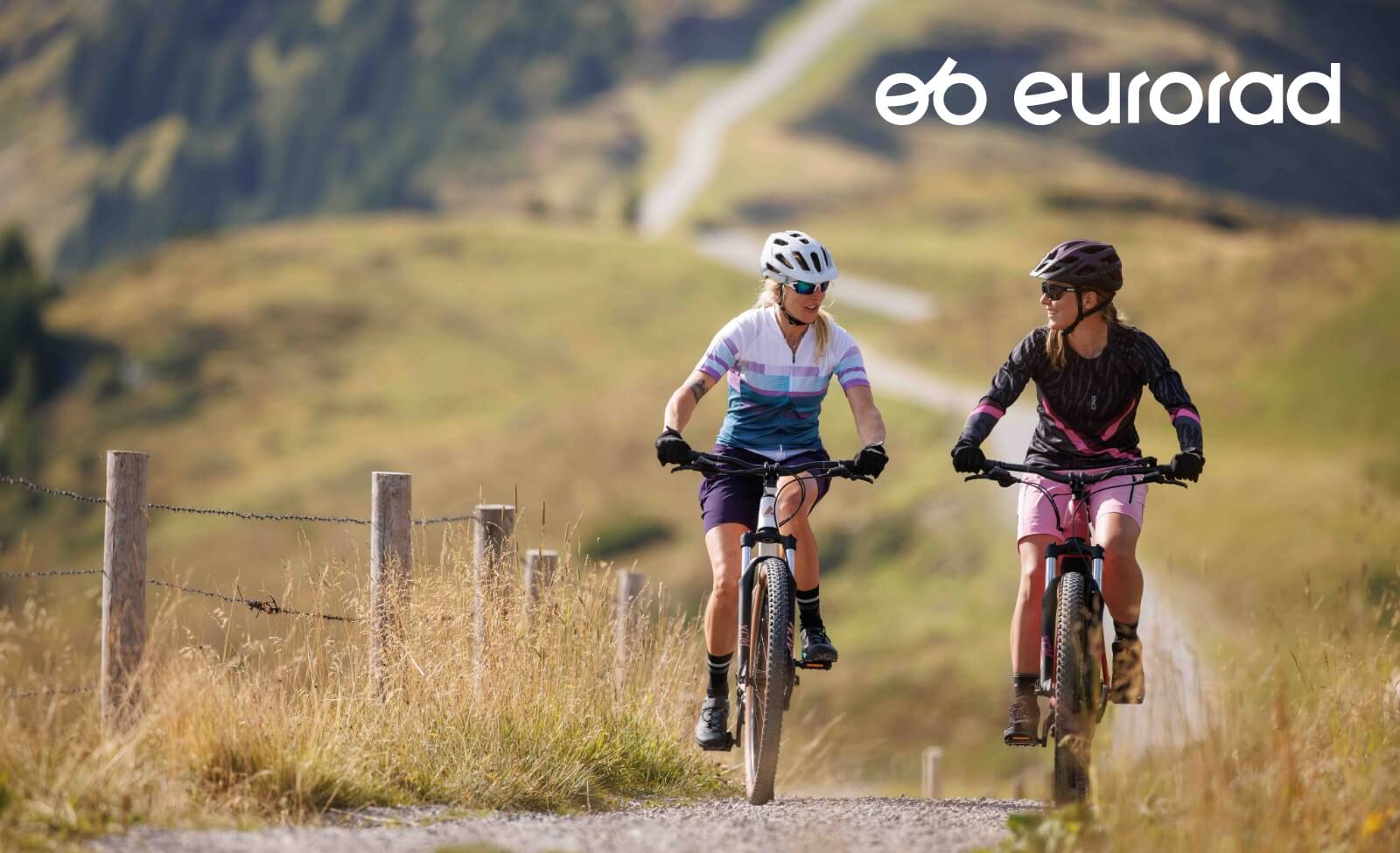 Zwei Frauen fahren mit Mountainbikes auf einem Schotterweg durch eine hügelige Landschaft. Logo: eurorad.