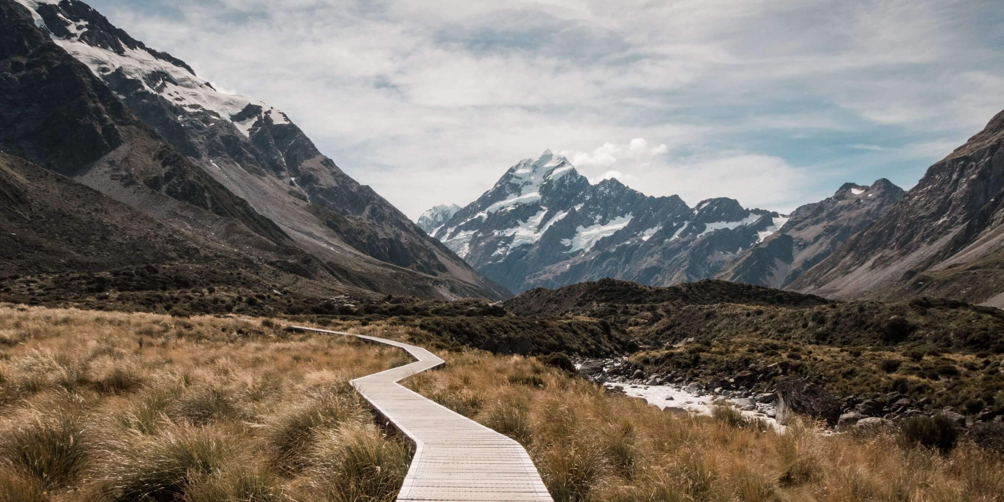 Holzweg durch alpine Berglandschaft mit schneebedeckten Gipfeln unter bewölktem Himmel. Ideal für Wanderer.