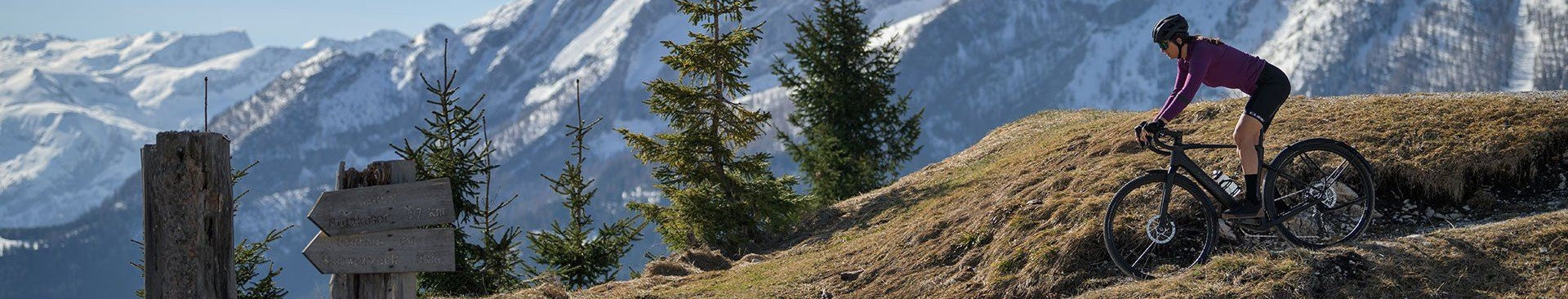 Radfahrer auf einem Cube Nuroad Hybrid Gravelbike in bergiger Landschaft mit schneebedeckten Gipfeln im Hintergrund.